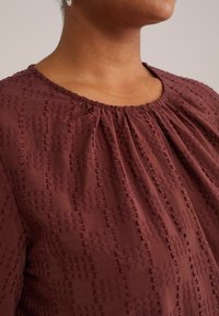 Blusa burdeos con un escote fruncido, que presenta un patrón texturizado de pequeños cuadrados y puntadas en una tela suave.