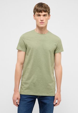 Junger Mann steht aufrecht, trägt ein hellgrünes T-Shirt mit kurzen Ärmeln und blaue Jeans vor einem schlichten hellen Hintergrund.