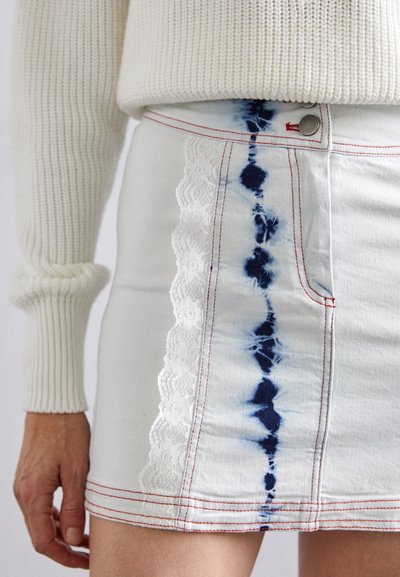 Mini-jupe en denim blanc avec un motif tie-dye bleu, des coutures rouges et une finition en dentelle blanche sur le côté. Bouton attaché à la taille.