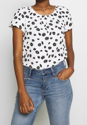 Mujer con blusa blanca de manga corta con estampado de flores negras y amarillas, combinada con vaqueros de mezclilla azul claro, una mano en el bolsillo.