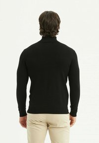 Pull à col roulé noir à manches longues et ourlet côtelé, mettant en valeur une texture tricotée lisse. Modèle de dos, associé à un pantalon beige.
