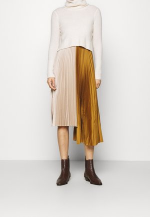 Kobieta w białym krótkim swetrze z golfem, dwukolorowej plisowanej spódnicy midi w beżu i musztardzie oraz brązowych botkach, stojąca na tle białej ściany.