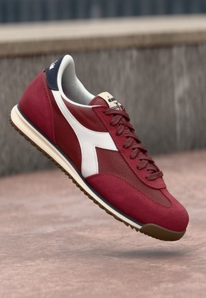 Chaussure de sport rouge avec une tige en daim et en mesh, détails en accent blanc, fermeture à lacets et semelle extérieure en caoutchouc gum.