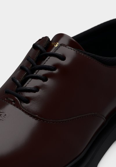 Κοντινό πλάνο γυαλιστερού σκούρου καφέ δερμάτινου παπουτσιού επίσημου τύπου με μαύρα κορδόνια και λεπτομερείς ραφές σε λευκό φόντο.