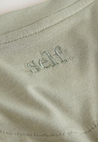 Beige stof med en nærbillede af det broderede ord "self." nær en søm, som viser tekstur og syningsdetaljer på beklædningen.