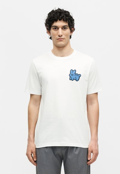 Άνδρας με σγουρά σκούρα μαλλιά που φοράει λευκό μπλουζάκι με μικρό γαλάζιο κουνελάκι στο αριστερό μέρος του στήθους και γκρι παντελόνι.