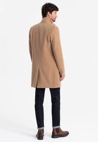 Beige ullkappa med hög krage, singelskärning bak, och knäppte ärmar, kombinerad med mörka jeans och bruna anklarstövlar.