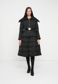 Black puffer coat with a high collar, gold zipper, and belt. Features a knee-length skirt with a pleated design and side pockets.