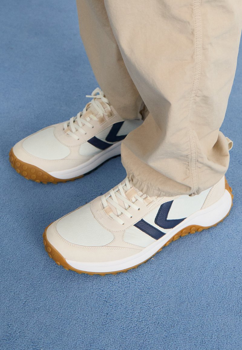 Helle beige Sportschuhe mit dunkelblauen Akzenten, texturiertem Obermaterial und Gummisohle. Schnürsenkel und gepolsterter Kragen für zusätzlichen Komfort.