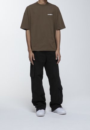 Homme portant un t-shirt marron oversize avec un petit texte blanc, un pantalon cargo noir avec des poches latérales et des baskets blanches sur un fond blanc uni.