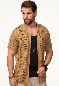 Strickjacke in Hellbraun mit Lochmuster, kurzen Ärmeln, einem Kragen und Knöpfen; über einem schwarzen Shirt mit Taschendetail getragen.