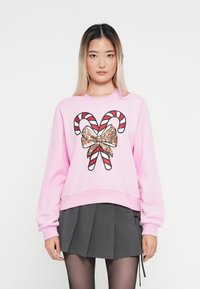 Różowy sweter z błyszczącą złotą kokardą i grafiką cukrowej laski, z długimi rękawami i luźnym krojem, połączony z ciemną plisowaną spódnicą.