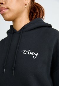 Črna kapuca iz mehkega blaga, ki ima na levem delu prsnega koša belo vezeno logotip "obey", z vrvicami in sproščenim krojem.
