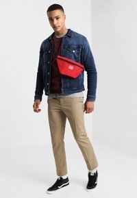 Red fabric fanny pack with a diagonal zipper and logo, worn over a blue denim jacket, paired with beige trousers and black sneakers.