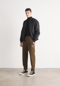 Schwarze Bomberjacke über einem schwarzen Rollkragenpullover, kombiniert mit braunen Jogginghosen mit Logo-Details. Dunkelblaue High-Top-Sneaker.