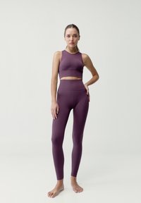 Fioletowy sportowy top z krótkim rękawem i dopasowanymi legginsami z wysokim stanem, z teksturą prążkowaną i bezszwowym designem. Modelka stoi boso.