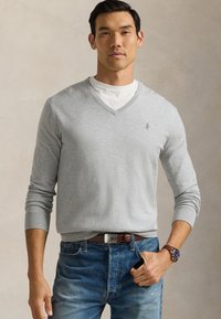 Sweater cinza claro em malha suave, usado sobre uma camisa branca, combinado com jeans azuis e um cinto castanho, apresentando um pequeno logotipo no peito.