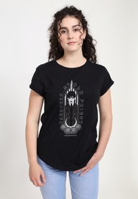 Mladá žena s kudrnatými vlasy, oblečená v černém tričku s goth-style ilustrací a textem "Temnota: Můj starý přítel", v kombinaci s světlými džíny.