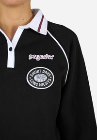 Μαύρο πόλο μπλούζα με λευκό γιακά, κόκκινη ρίγα και κεντητό λογότυπο. Διαθέτει πατιλέτα με την επιγραφή "ΚΟΙΝΩΣ ΜΕΡΕΣ ΜΕΓΑΛΕΣ ΝΥΧΤΕΣ" στο στήθος. Μαλακή υφή.