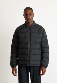 Schwarze Daunenjacke mit Reißverschluss vorne, gerippten Bündchen und Stehkragen. Mit horizontalem gestepptem Design und dezentem Branding.