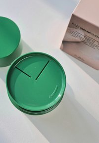 Zielony okrągły pojemnik o gładkiej powierzchni, z dwoma szczelinami na pokrywie i detalem w kształcie strzałki, umieszczony obok pudełka produktu.