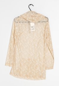Blusa trasparente beige con maniche lunghe, ricami floreali bianchi, cappuccio e orlo curvo. Etichetta di cura attaccata visibile sul retro.