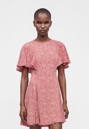 Femme aux cheveux courts portant une courte robe rouge et blanche à pois avec des manches volantes, debout sur un fond uni.