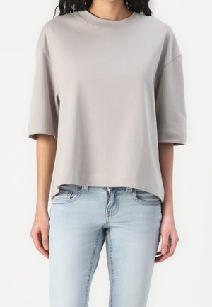 Frau trägt ein locker sitzendes, hellgraues T-Shirt mit kurzen Ärmeln und hellblaue Jeans, steht vor einem schlichten weißen Hintergrund.