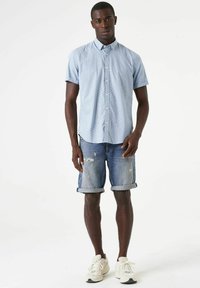 Lichtblauw shirt met korte mouwen en een geometrisch patroon, gecombineerd met versleten spijkerbroekshorts en witte sneakers. Platte kraag en knoopsluiting.