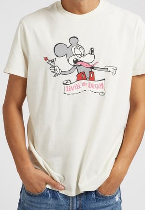 Kremowa bawełniana koszulka z króliczkiem trzymającym koktajl, w czerwonych szortach i z banerem, na którym widnieje tekst "Żyjąc marzeniem" w kolorze różowym.