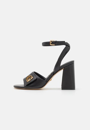 Sandal in pelle nera con tacco alto, dotata di una fascia ampia, decorazione con logo dorato e una fibbia strutturata alla caviglia. Tacco a blocco quadrato.