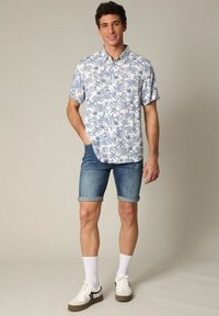 Korte mouwen, knoopshirt met een witte achtergrond en een blauw palmbladpatroon, gecombineerd met opgerolde denimshorts en witte sneakers.