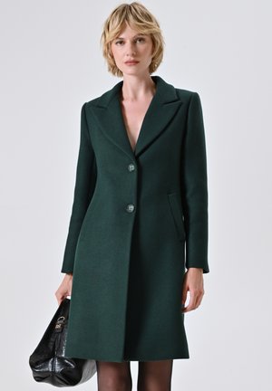 Cappotto verde di lana con revers a sciallata, due bottoni e tasche laterali. Il modello tiene una borsa nera, in piedi davanti a uno sfondo neutro.