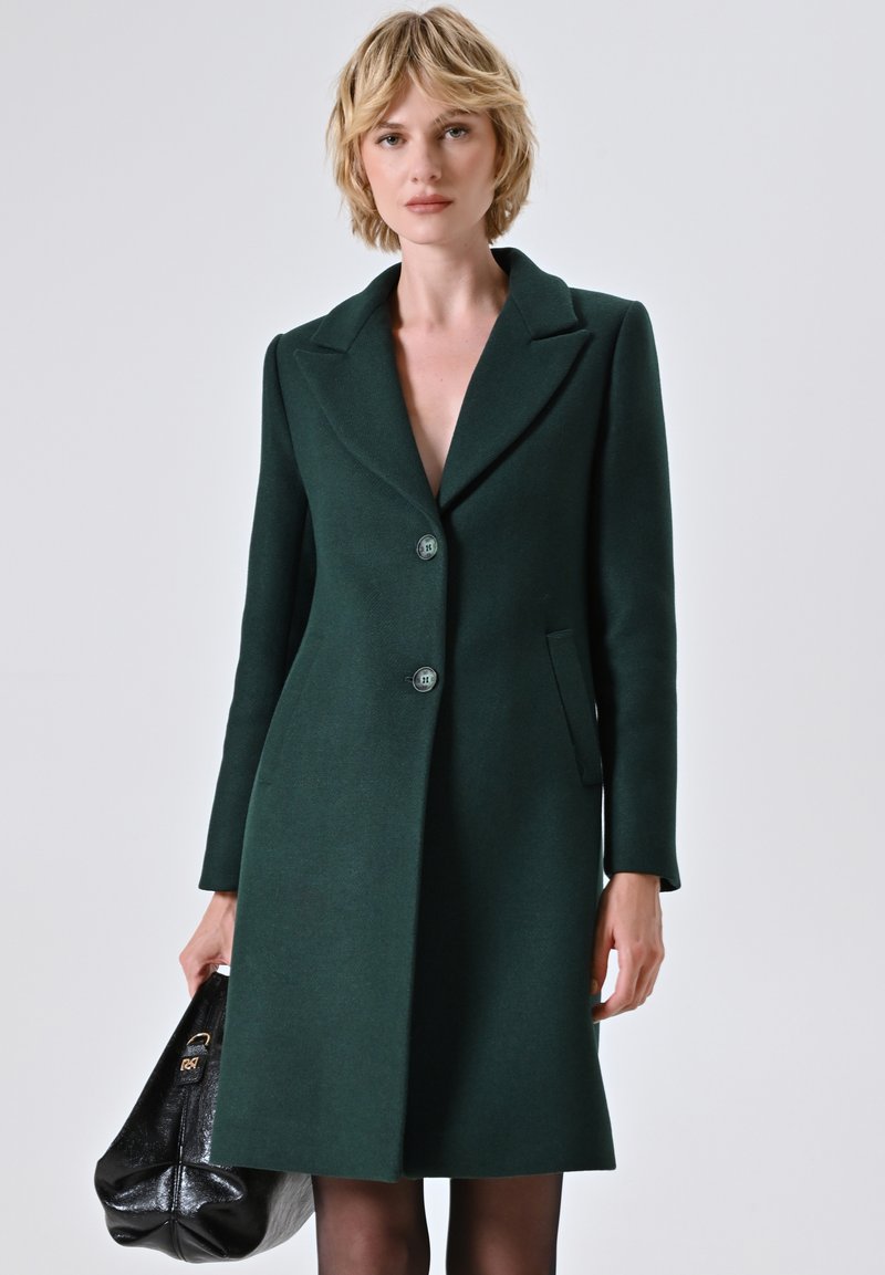 Cappotto verde di lana con revers a sciallata, due bottoni e tasche laterali. Il modello tiene una borsa nera, in piedi davanti a uno sfondo neutro.