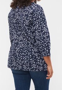 Top floreale navy con piccolo motivo a fiori bianchi, maniche a tre quarti, materiale morbido, orlo arrotondato, abbinato a jeans scuri blu.