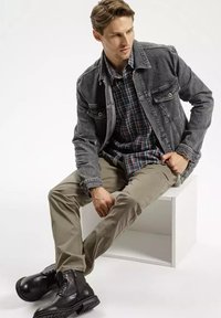 Szary dżinsowy kurtka nałożona na koszulę w kratę, beżowe spodnie i czarne buty na sznurówki. Gładkie tekstury z wyraźnymi wzorami w odzieży.