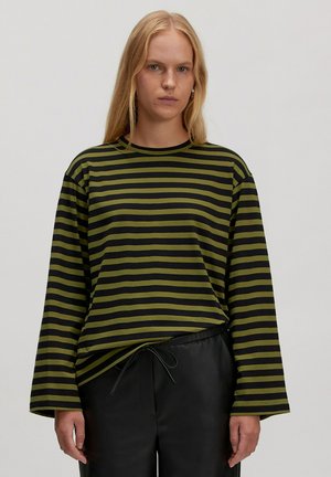 Junge Frau mit langen blonden Haaren, die ein schwarz-olivgrün gestreiftes Langarmshirt und schwarze Hosen trägt, vor einem einfarbigen Hintergrund.