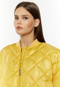 Giacca bomber imbottita gialla con finitura lucida, dotata di zip anteriore e colletto a costine. La texture è liscia con un motivo geometrico.