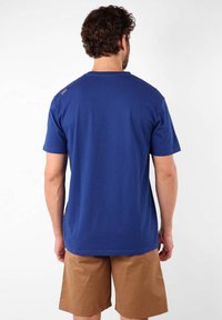 T-shirt bleu à manches courtes en coton, avec un col rond et un petit détail de logo sur l'épaule, associé à un short beige.