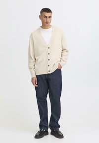 Ung mann står selvsikkert i en kremfarget cardigan over en hvit skjorte og mørkeblå jeans, med kort hår og svarte sko.