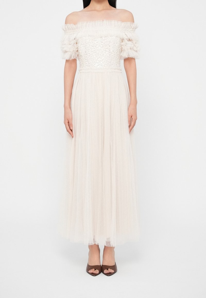 Femme portant une robe blanche épaules dénudées avec un corsage texturé et des manches bouffantes transparentes en couches, associée à des talons ouverts marron.