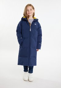 Lunga giacca piumino blu navy con foderina gialla sulla cappuccio, zip completa, tasche e dettaglio del logo sul petto. Abbinata a jeans e stivali bianchi.
