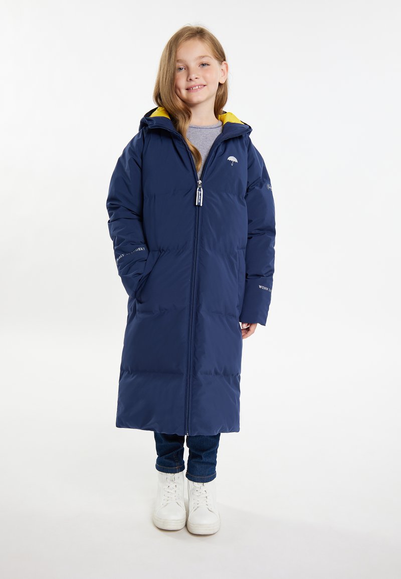 Lunga giacca piumino blu navy con foderina gialla sulla cappuccio, zip completa, tasche e dettaglio del logo sul petto. Abbinata a jeans e stivali bianchi.