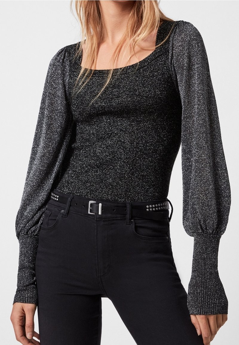 Haut ajusté noir et argenté scintillant à manches longues avec manches bouffantes et larges poignets côtelés, associé à un pantalon noir et une ceinture cloutée.