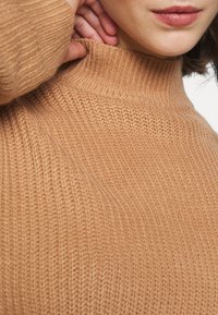 Pletený rolák v teplé barvě tanu s žebrovanou texturou, s vyšším límcem a přiléhavými rukávy.