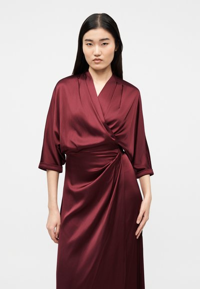 Γυναίκα με μακριά σκουρόχρωμα μαλλιά που φορά ένα βαθύ κόκκινο σατέν φόρεμα τύπου μανδύα με μανίκια τριών τετάρτων, μπροστά σε απλό φωτεινό φόντο.