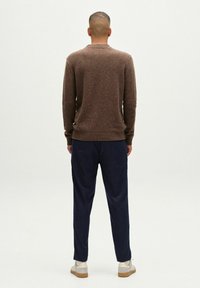 Brązowy sweter dzianinowy z prążkowanymi mankietami i dołem, zestawiony z ciemnoniebieskimi spodniami. Model nosi jasne buty. Tło jest gładkie, białe.