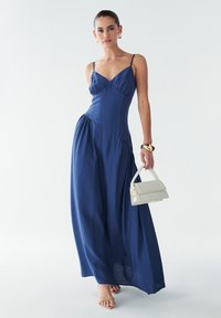 Tumši zila maksi kleita ar smalkām spilventiņām, piegulošu korseti un savākto vidukli. Valkāta ar gaišas krāsas roku somu un bēšām sandālēm.