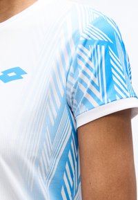 Κοντινό πλάνο του άνω βραχίονα ενός ατόμου που φοράει λευκό και μπλε αθλητικό πουκάμισο με γεωμετρικά σχέδια και μπλε λογότυπο στο στήθος.