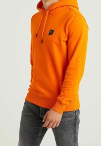 Uomo che indossa un felpa arancione brillante con cappuccio e piccoli lacci, accompagnata da un piccolo logo nero sul petto, abbinata a jeans neri sbiaditi.